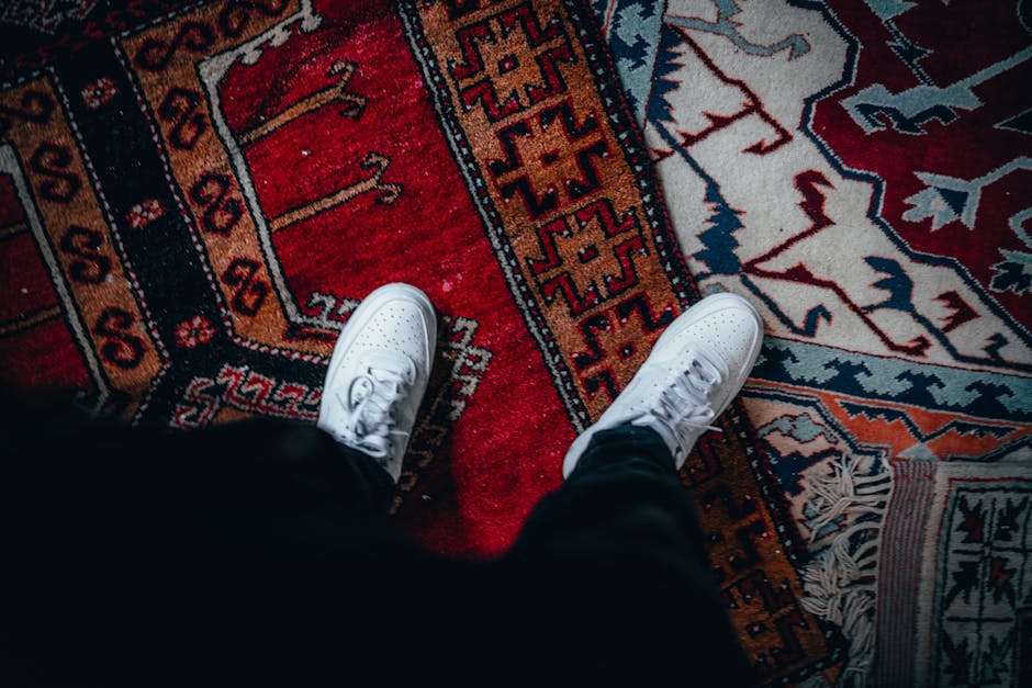A top-down view of a person's white sneakers standing on a colorful, patterned area rug with intricate geometric and floral designs, predominantly in red, navy, and beige tones. The rug appears clean 