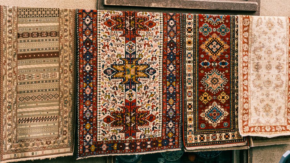 A top-down view of a person's white sneakers standing on a colorful, patterned area rug with intricate geometric and floral designs, predominantly in red, navy, and beige tones. The rug appears clean 