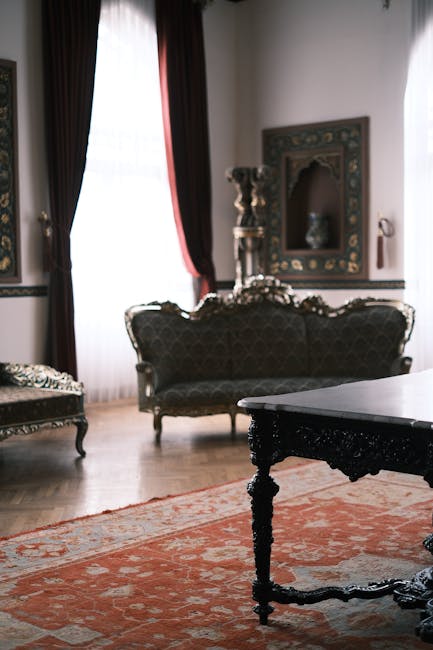 A person wearing black gloves is using a handheld electric fabric steamer to deep clean and sanitise a dark grey upholstered sofa in a living room. The sofa features a modern design with wide armrests